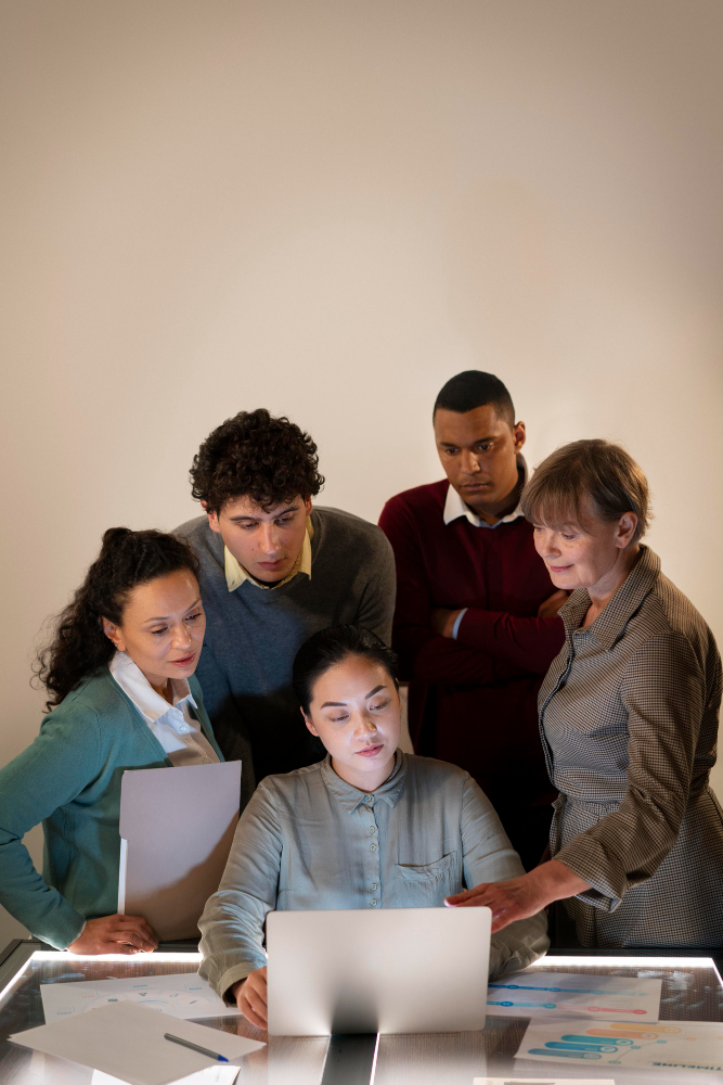 Five colleagues gathered around a laptop at a lit glass table